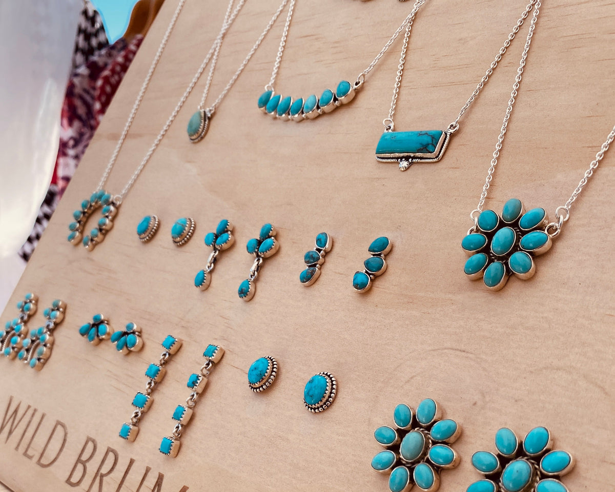 Turquoise jewelry displayed on a wooden sign engraved with the brand name 'WILD BRUMBY CO.' The jewelry includes necklaces and earrings with bright turquoise stones set in silver-toned metal, arranged on a woven surface.